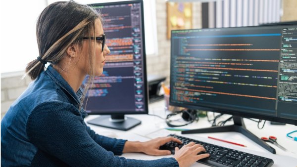 Photo of a woman working on computer code at two monitor screens. 